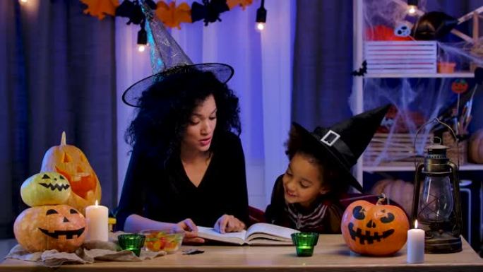 穿着节日服装和巫婆帽子的非洲裔美国小女孩看书。妈妈和女儿坐在万圣节之夜装饰的房间的桌子旁。慢动作