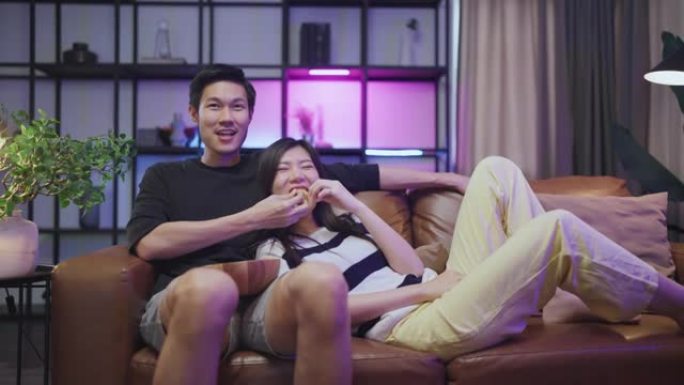 娶成人亚洲人在家看电视，亚洲夫妇欢呼体育比赛与笑笑胜利沙发沙发在客厅家庭隔离活动