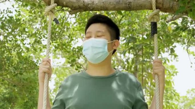 亚洲男孩戴着口罩在公园户外的树下玩绳子秋千，新的正常生活方式概念。