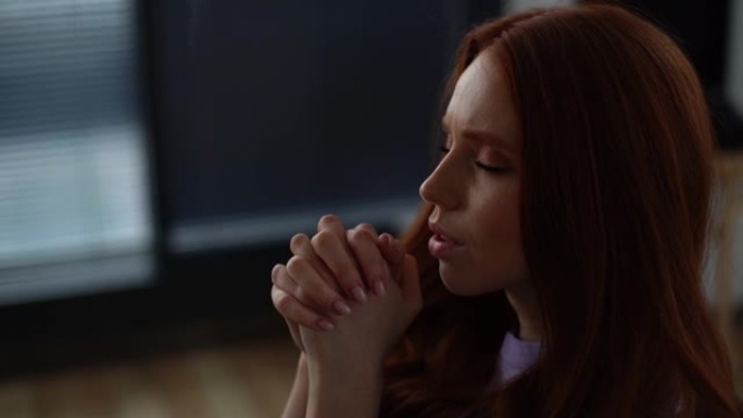 在黑暗的客厅里，沮丧的年轻女子闭着眼睛祈祷的特写镜头，双手交叉在脸前。