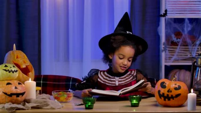 穿着节日服装的非洲裔美国小女孩热情地看书。孩子坐在万圣节之夜装饰的房间的桌子旁。和家人一起度过可怕而