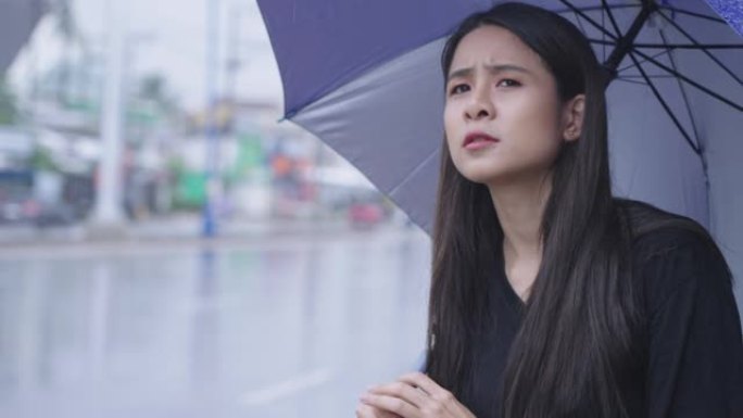 一个美丽的东亚妇女独自站在路边等待出租车或公共汽车，在下雨的时候，在雨中撑伞，在高峰时间，热带气候变