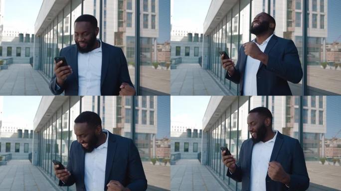 穿着正式西装的快乐兴奋的大胡子非洲男子使用手机，用手示意是手势，从积极的情绪中惊呼，庆祝胜利，站在商