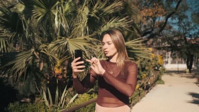 迷人的时尚女孩使用智能手机在美丽的公园散步时与朋友视频聊天。美丽的女孩切换到前置摄像头聊天以显示视图