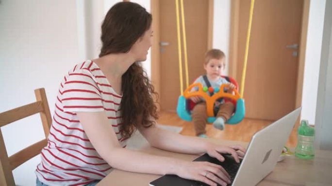 开朗的母亲在家里的办公室里用笔记本电脑工作时摇摆着男婴