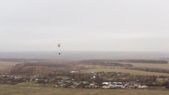 穿着斗篷的人像超人一样在滑翔伞上飞过黄色的秋天。行动。雾天在村庄附近飞行跳伞者。