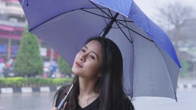 一名年轻美丽的亚洲妇女站在路边，在热带台风引起的雨天手持蓝色雨伞，在城市路边等公共汽车，天气预报，感
