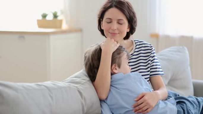 快乐的母亲拥抱儿子跪下睡觉。可爱的高加索妈妈和学龄前儿童在家沙发上拥抱