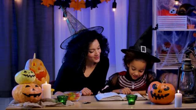 穿着节日服装和巫婆帽子的非洲裔美国小女孩看书。妈妈和女儿坐在万圣节之夜装饰的房间的桌子旁。慢动作