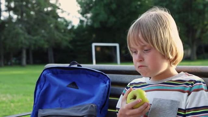 小学生背着背包坐在学校院子附近的长凳上，放学后或课间休息时吃苹果作为零食