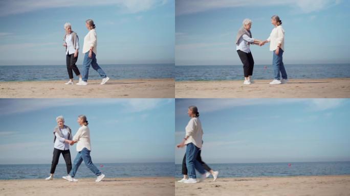 追踪两名开朗自发的高级妇女在沙滩上牵着手行走，快乐地交谈和牵着手奔跑的宽镜头。快乐的退休女友喜欢在一