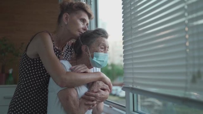 成熟的女儿去疗养院看望老妈。由于冠状病毒感染，年迈的女儿站在窗户上时，戴着医用口罩拥抱着她90岁的母