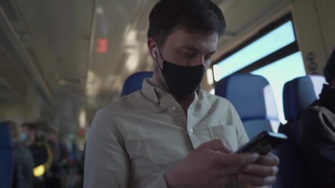 戴着口罩和耳机的男子乘客在乘火车安全旅行时使用电话在线观看视频和阅读新闻。公共交通工具中的病毒保护和