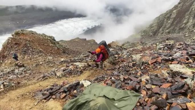 一位精疲力竭的游客背着大背包，沿着山间小路跌跌撞撞地走过石头和巨石