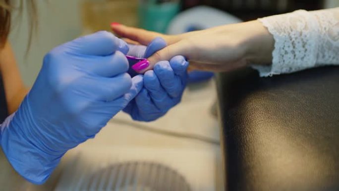 戴着手套的面目全非的人在沙龙画年轻女子的指甲。未知的美甲师在美容院为客户提供指甲油修指甲。护理和美容