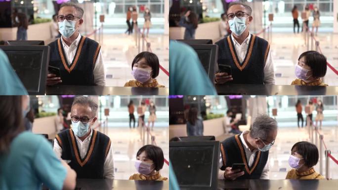戴着口罩的亚洲华人祖父孙女在电影院售票处使用手机应用程序购买电影票