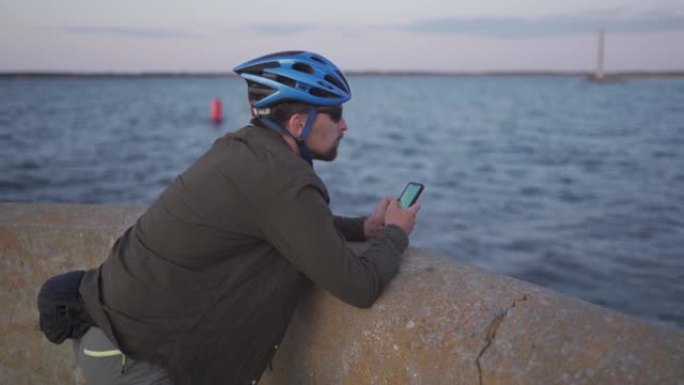 日落时的海上自行车旅行。男性骑自行车的人在海上的路堤上休息，并使用智能手机建造路线并导航GPS。在海