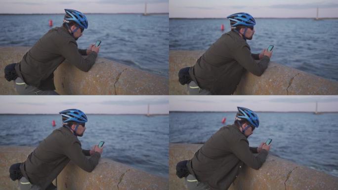 日落时的海上自行车旅行。男性骑自行车的人在海上的路堤上休息，并使用智能手机建造路线并导航GPS。在海