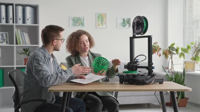 女性和男性工程师使用现代技术，并在手讨论3D模型，同时在房间里坐在桌子上的3D打印机上工作
