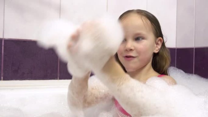 漂亮的女孩穿着粉红色的泳衣，浴缸里有很多泡沫。她玩着手上的泡沫。
