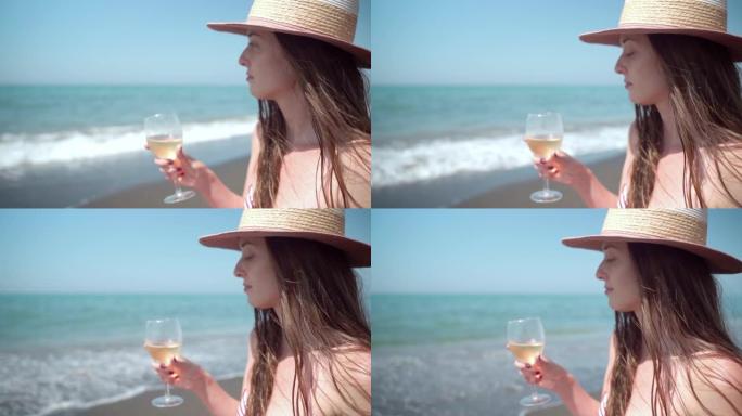 在令人惊叹的海浪背景下，戴着夏日帽子的思考女人手里拿着一杯新鲜的葡萄酒，放松的女性在海边享受暑假。炎