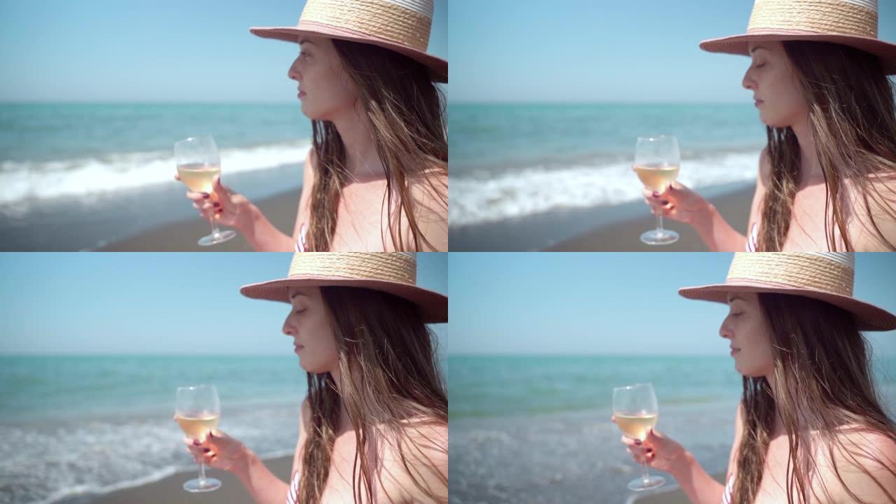 在令人惊叹的海浪背景下，戴着夏日帽子的思考女人手里拿着一杯新鲜的葡萄酒，放松的女性在海边享受暑假。炎