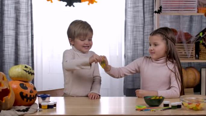 在万圣节装饰的房间里，一个男孩和一个女孩坐在一张桌子旁，桌子上放着零食和南瓜。孩子们玩果冻虫。万圣节
