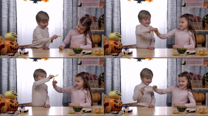 在万圣节装饰的房间里，一个男孩和一个女孩坐在一张桌子旁，桌子上放着零食和南瓜。孩子们玩果冻虫。万圣节