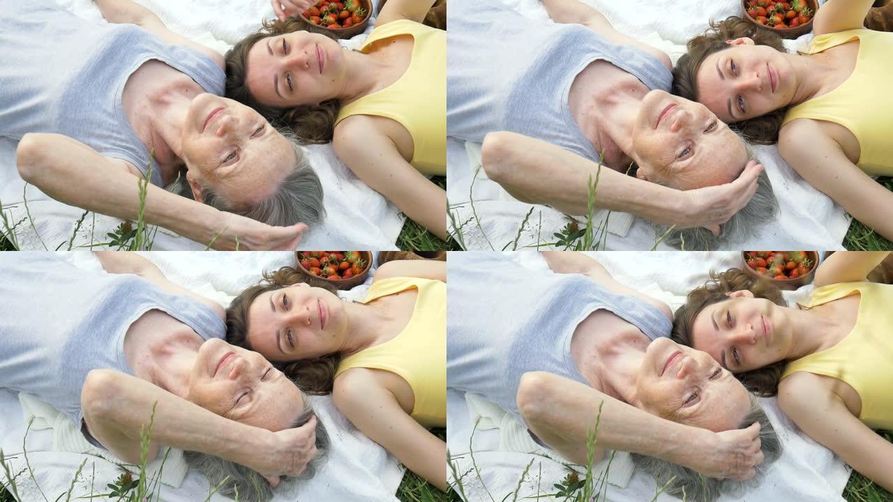 美丽的老祖母，白发和皱纹的脸正躺在白色地毯上，她的成年女儿在户外野餐，母亲节，退休快乐