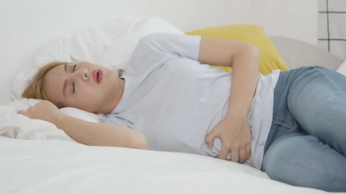 亚洲女人穿着上腹部腹痛，躺在床上，阳光直射，躺在白色卧室里。