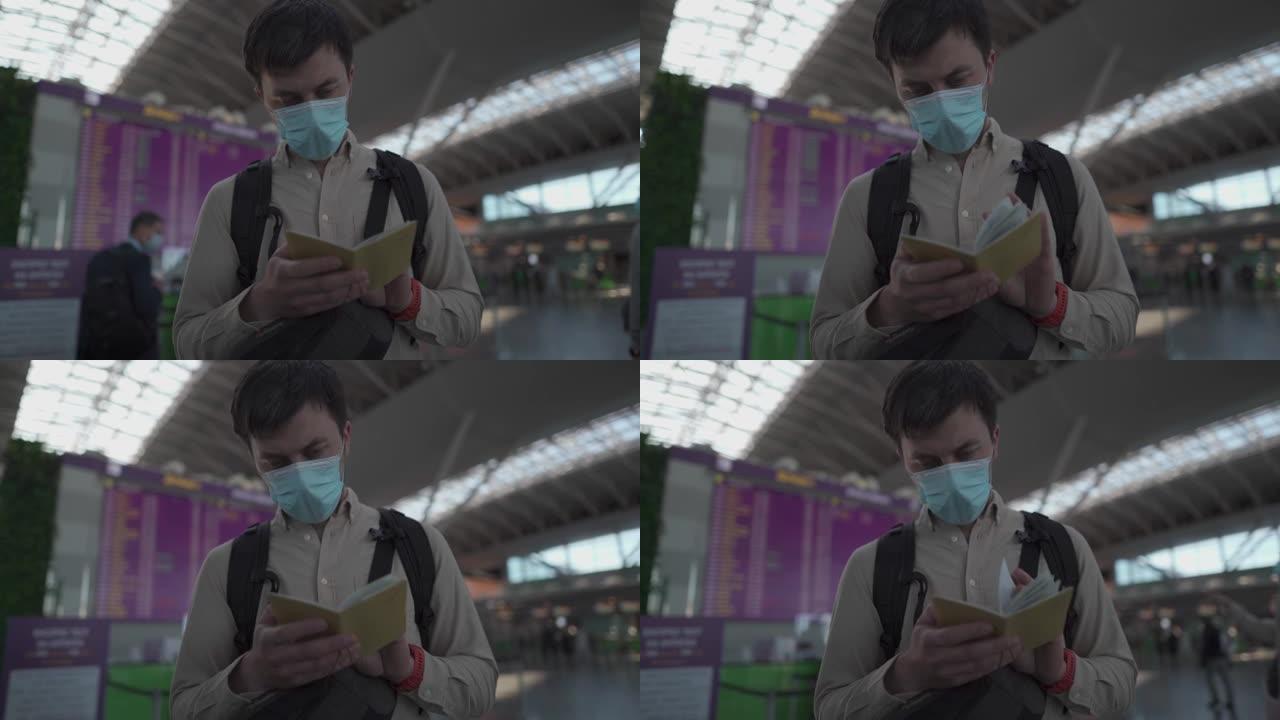 在机场戴防护面具的男子在电子时刻表附近持有护照。在冠状病毒大流行期间开放边境旅行。男性在机场旅行时检