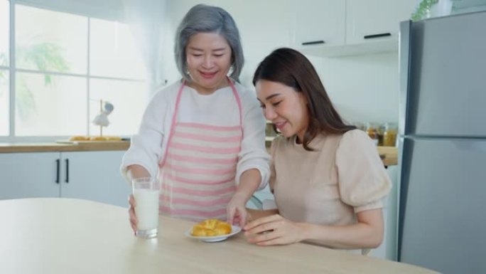 亚洲爱心老年母亲在厨房为女儿提供食物。美丽成熟的老妇人早上在家递羊角面包和牛奶来照顾年轻女孩。家庭活