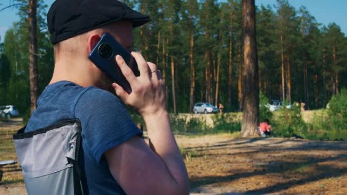 一个戴着帽子、留着胡子度假的年轻人正在用智能手机聊天。暑假在森林里治愈身体和灵魂。其他度假者也在安静
