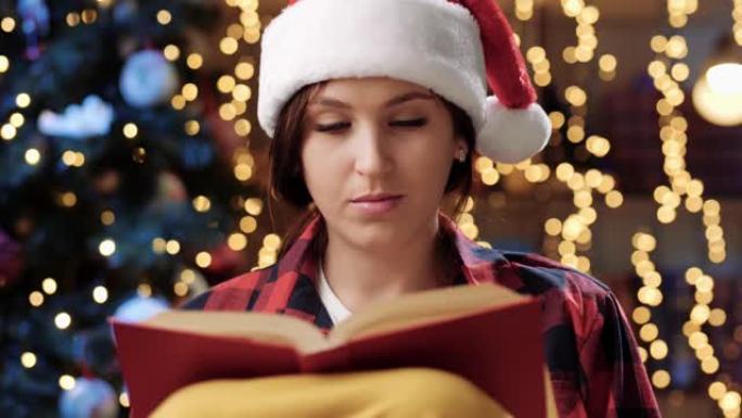 圣诞概念。戴着红帽子的漂亮女人的前视图坐在扶手椅上看书，喝杯子里的东西，背景是花环和圣诞树的模糊灯光