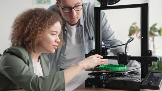 微笑的女人和一个男人使用现代技术，坐在桌子旁高兴地看着家里的3D打印机