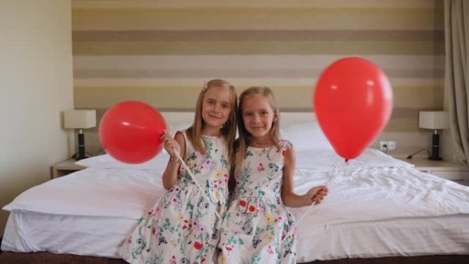 两个双胞胎姐妹坐在卧室的床上，上面放着红色的气球。