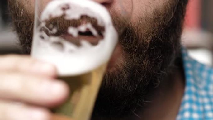 大胡子的人正在喝啤酒。男性的手拿着装满泡沫的玻璃杯到嘴里，喝啤酒。特写
