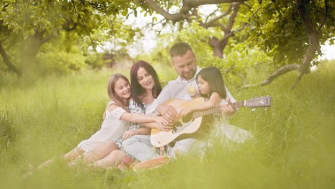 幸福的家庭，可爱的女儿在绿色花园野餐，吉他歌曲。父亲弹吉他，母亲拥抱可爱的女儿。男人为妻子和女儿弹吉