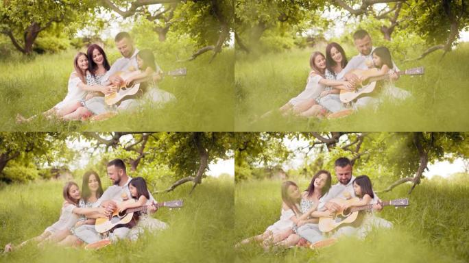 幸福的家庭，可爱的女儿在绿色花园野餐，吉他歌曲。父亲弹吉他，母亲拥抱可爱的女儿。男人为妻子和女儿弹吉