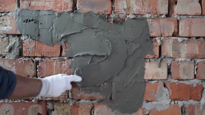 他戴着手套的大师级建筑工人的手的特写镜头，拿着刮刀并用灰泥涂抹在砖墙上。