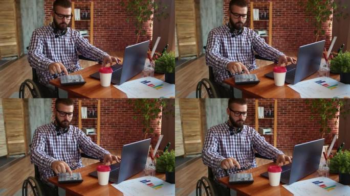 戴着眼镜的轮椅商人在笔记本电脑上工作，使用带桌子的桌子上的计算器，侧视图。残疾人使用数字小工具的概念