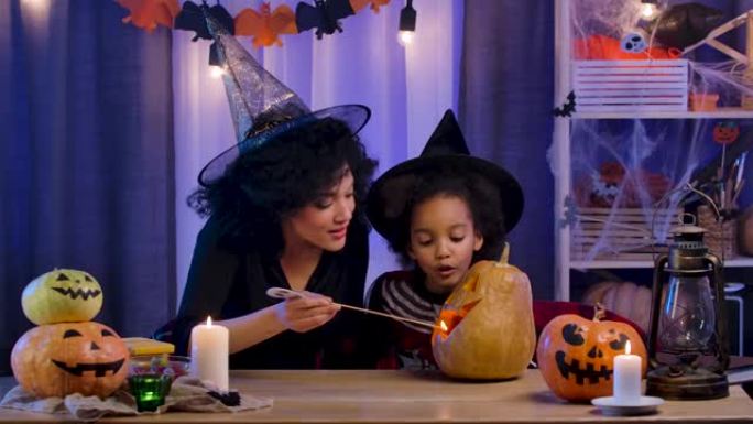 小女孩和女人，戴着巫婆帽的非洲裔美国人，用雕刻的笑脸点燃南瓜蜡烛。妈妈和女儿坐在万圣节之夜装饰的房间
