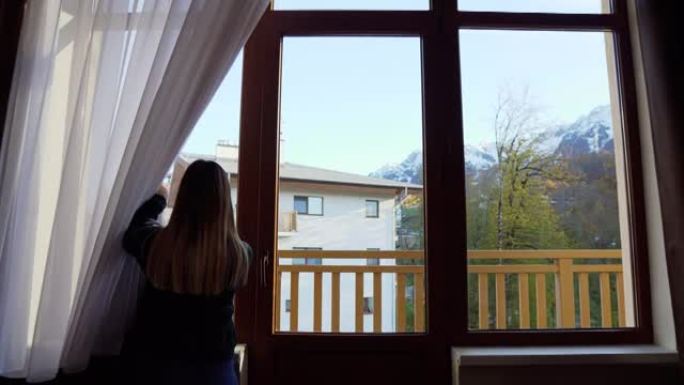 女孩将窗帘从窗户推开，窗外可见凉爽的山景。摄像机移动到窗口