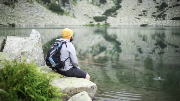 男性游客坐在湖边的一块石头上，将石头掉入水中