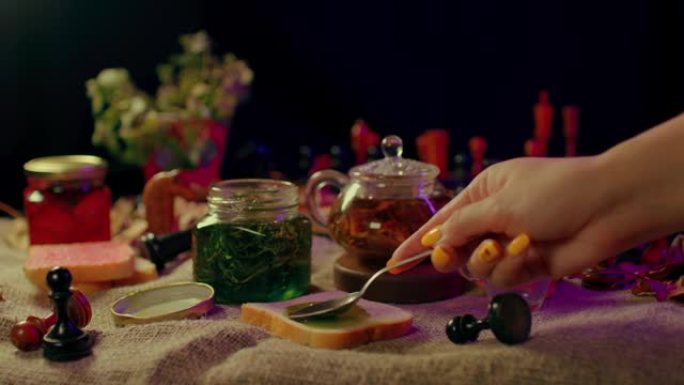 桌子上放着果酱和一块面包的小罐子的特写镜头。无法辨认的女人用茶匙在吐司上撒上果酱