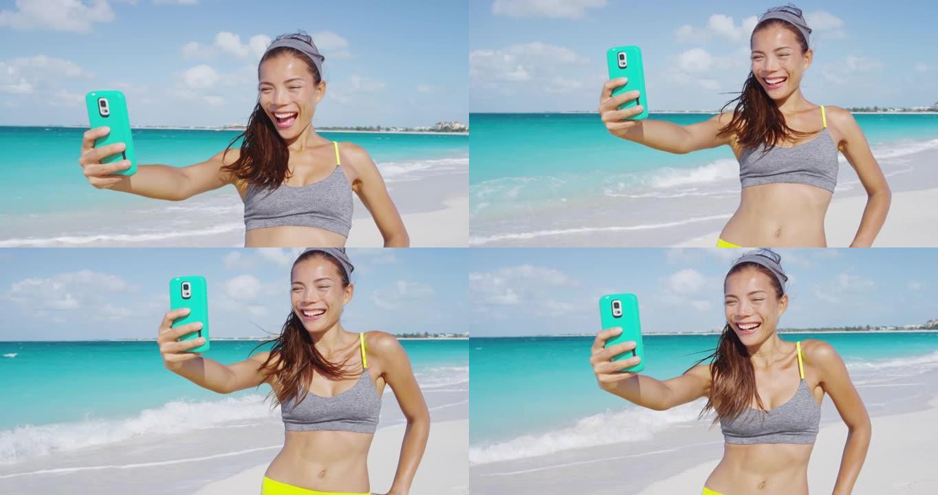 快乐的运动女孩在沙滩上跑步后笑着用手机自拍。女人在暑假锻炼后进行健身自拍，过着健康的生活方式。红色慢