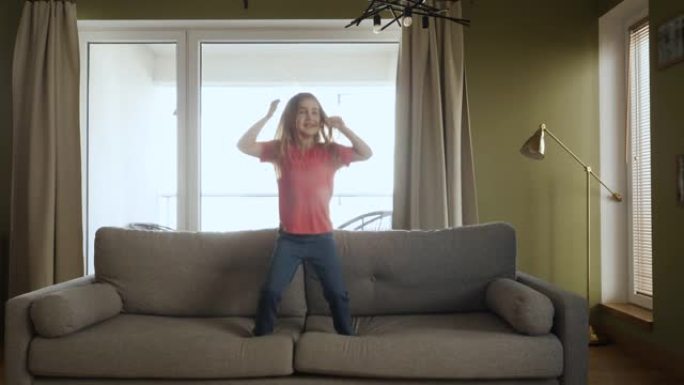 有趣快乐的小女孩正在家里的沙发上跳舞和跳跃。女儿在客厅玩耍。热爱生活方式之家。快乐的小女孩在沙发上随