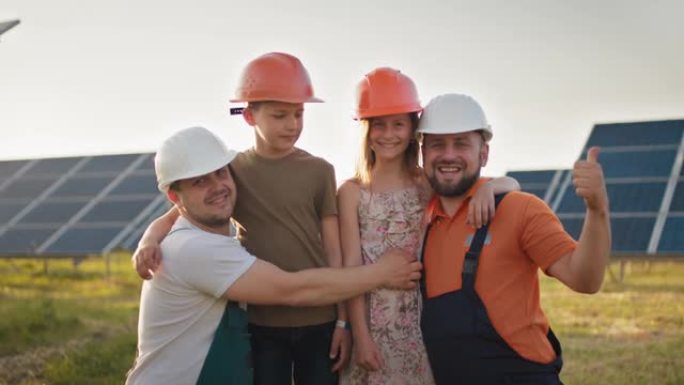 在太阳能发电厂，两个爸爸和两个戴着防护头盔的孩子的肖像，都指着相机并微笑着。父母在太阳能发电厂将头盔