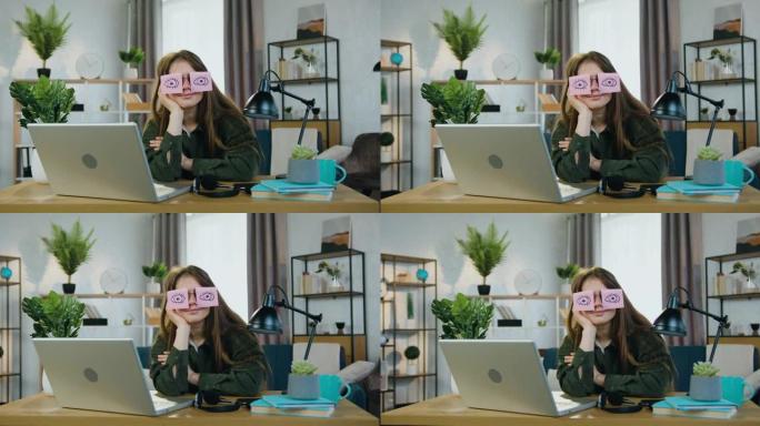 迷人有趣的年轻现代女性的肖像，她坐在家里的工作场所，戴着眼镜在相机上摆姿势，上面贴着贴纸，睁开眼睛
