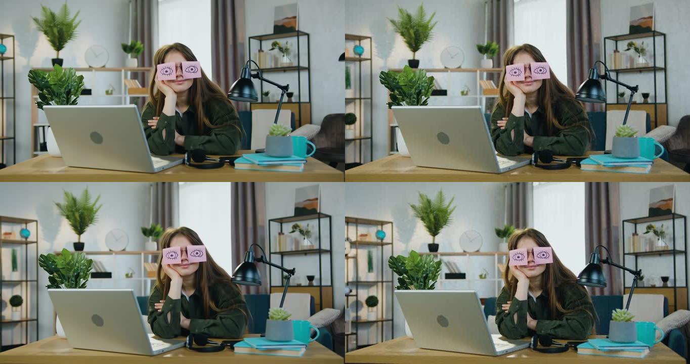 迷人有趣的年轻现代女性的肖像，她坐在家里的工作场所，戴着眼镜在相机上摆姿势，上面贴着贴纸，睁开眼睛